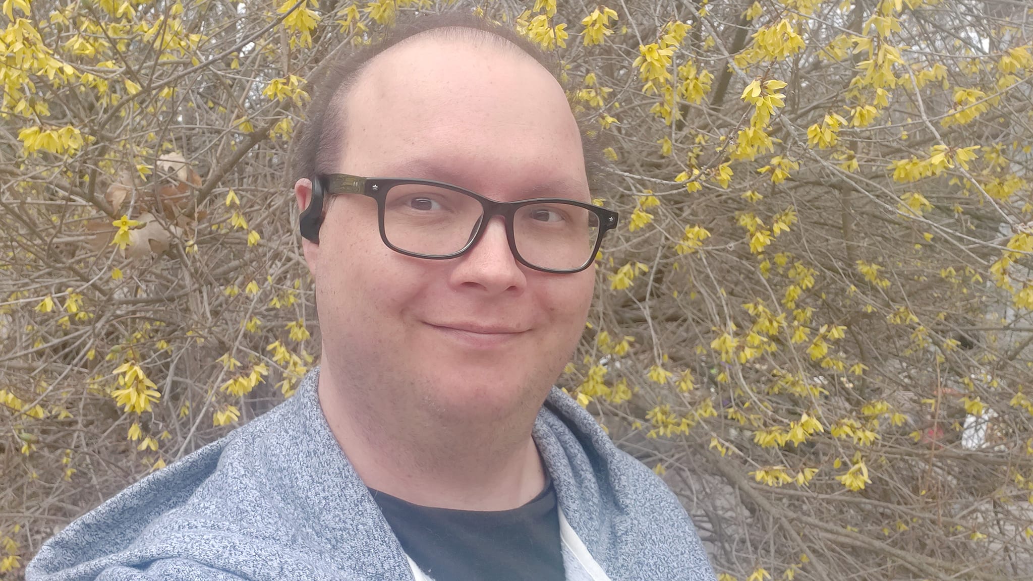 A man wearing a hoodie with the hood down stands in front of a bush with some yellow leaves